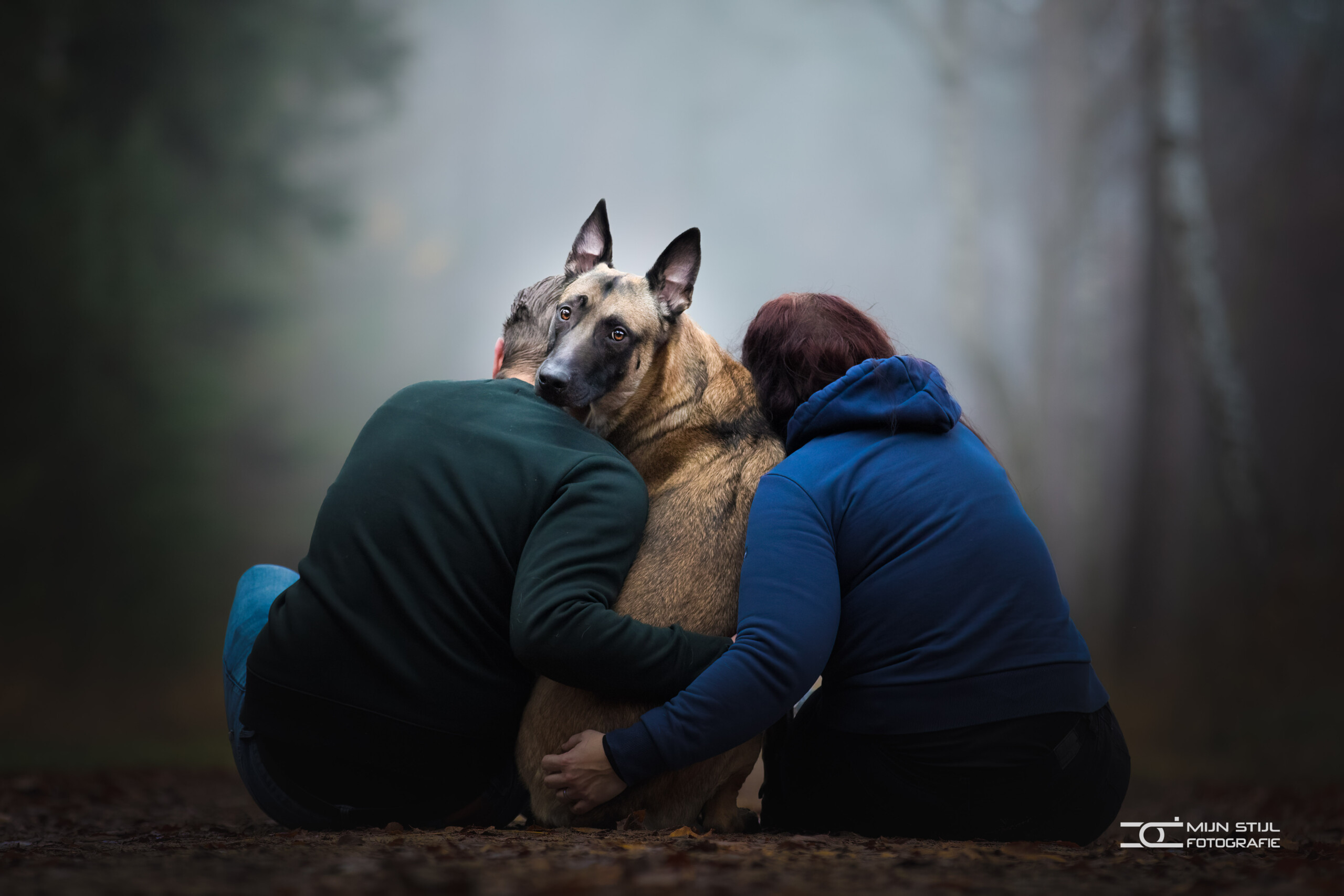 Een Mechelse herder tussen zijn twee baasjes, die hem samen omarmen in het bos